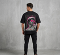 "Rear view of a male model wearing a black Akido oversized streetwear t-shirt featuring a large graphic print on the back. The graphic displays a grayscale statue of Michelangelo's David wearing vibrant neon pink sunglasses, surrounded by white graffiti text, doodles, and a pink neon outline, set against a concrete wall backdrop."