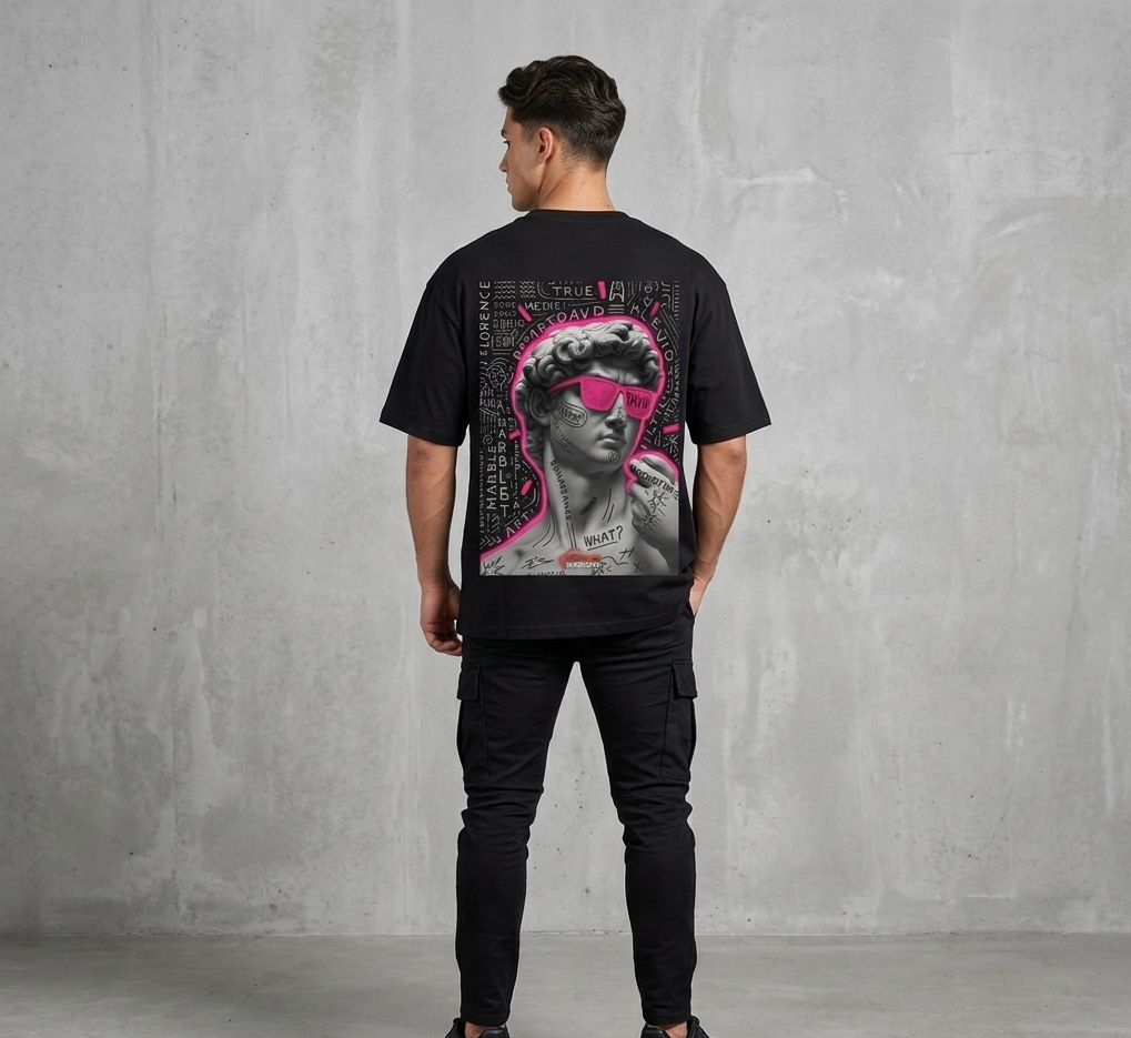 "Rear view of a male model wearing a black Akido oversized streetwear t-shirt featuring a large graphic print on the back. The graphic displays a grayscale statue of Michelangelo's David wearing vibrant neon pink sunglasses, surrounded by white graffiti text, doodles, and a pink neon outline, set against a concrete wall backdrop."