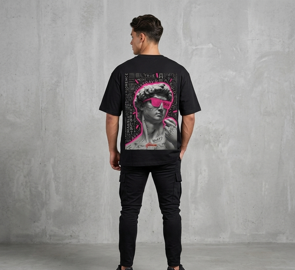 "Rear view of a male model wearing a black Akido oversized streetwear t-shirt featuring a large graphic print on the back. The graphic displays a grayscale statue of Michelangelo's David wearing vibrant neon pink sunglasses, surrounded by white graffiti text, doodles, and a pink neon outline, set against a concrete wall backdrop."