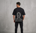 Man wearing an oversized black t-shirt with a white abstract line-art face graphic printed on the back, paired with black cargo pants against a gray concrete wall background.