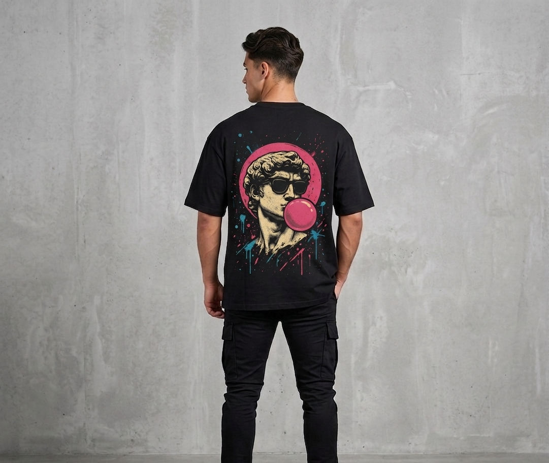 “Man wearing a black oversized Akido graphic t-shirt featuring a retro-style statue illustration with sunglasses and pink bubble gum artwork on the back, standing against a gray concrete wall.”