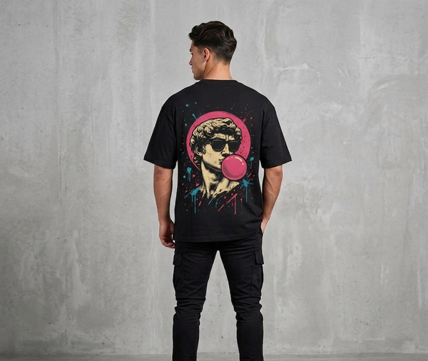 “Man wearing a black oversized Akido graphic t-shirt featuring a retro-style statue illustration with sunglasses and pink bubble gum artwork on the back, standing against a gray concrete wall.”
